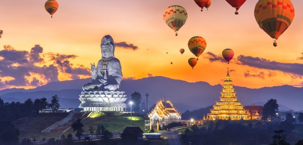 Sunset over Chiang Rai with a giant white Buddha statue and golden temple, surrounded by colorful hot air balloons in a serene, majestic landscape.
