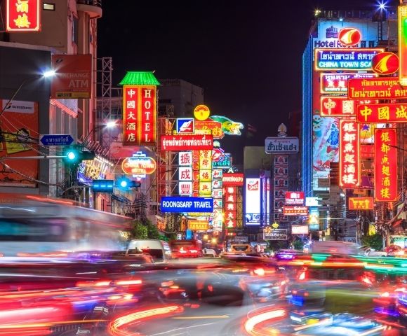A vibrant city street illuminated by colorful neon signs in various languages, bustling with blurred car lights and activity, exuding a dynamic nightlife energy.