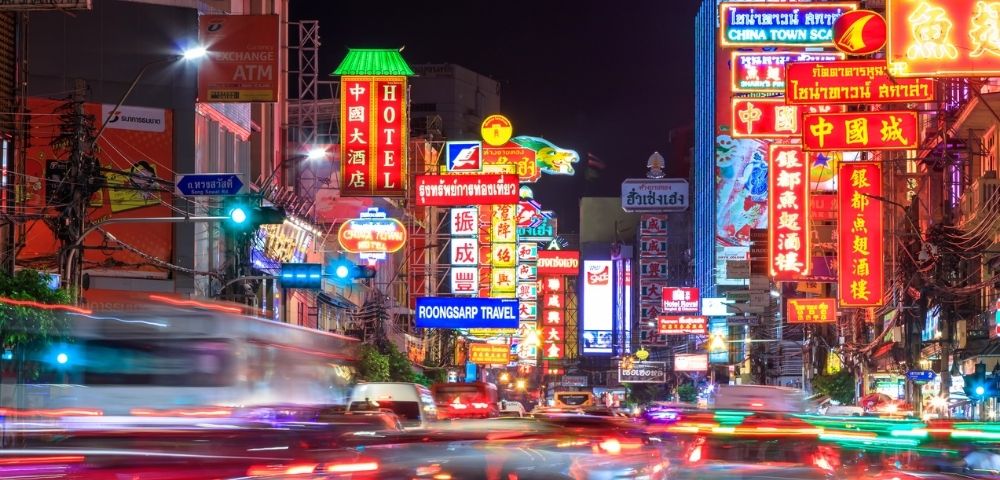A vibrant city street illuminated by colorful neon signs in various languages, bustling with blurred car lights and activity, exuding a dynamic nightlife energy.