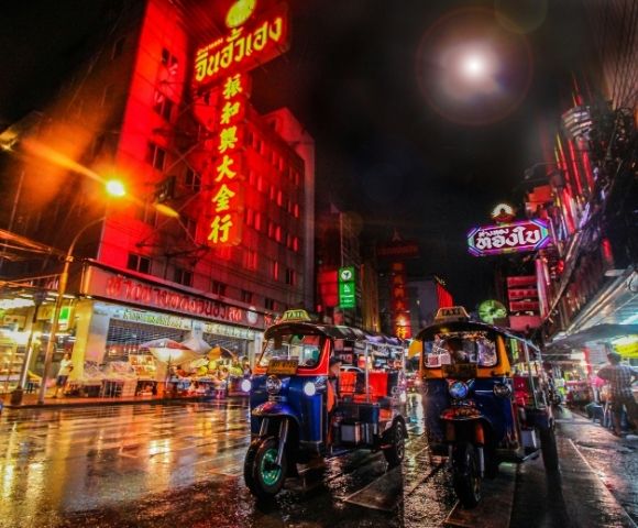 Bustling nighttime street in Bangkok with vibrant neon signs lighting up the scene. Tuk-tuks line the wet road, reflecting colorful lights.