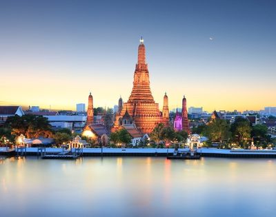 A serene view of Wat Arun temple in Bangkok at sunset, illuminated in warm tones. The Chao Phraya River reflects the colorful sky, creating a tranquil ambiance.