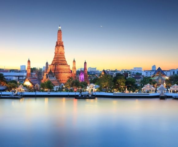 A serene view of Wat Arun temple in Bangkok at sunset, illuminated in warm tones. The Chao Phraya River reflects the colorful sky, creating a tranquil ambiance.