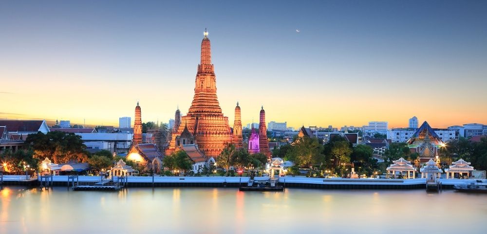 A serene view of Wat Arun temple in Bangkok at sunset, illuminated in warm tones. The Chao Phraya River reflects the colorful sky, creating a tranquil ambiance.