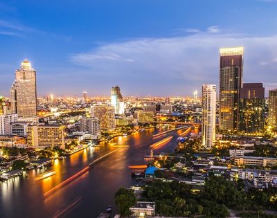 A vibrant cityscape at dusk with illuminated skyscrapers, bustling river traffic, and a twilight sky. The scene conveys energy and urban life.