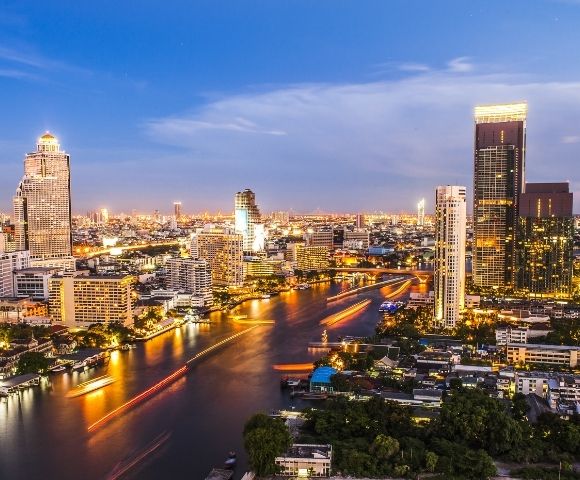 A vibrant cityscape at dusk with illuminated skyscrapers, bustling river traffic, and a twilight sky. The scene conveys energy and urban life.