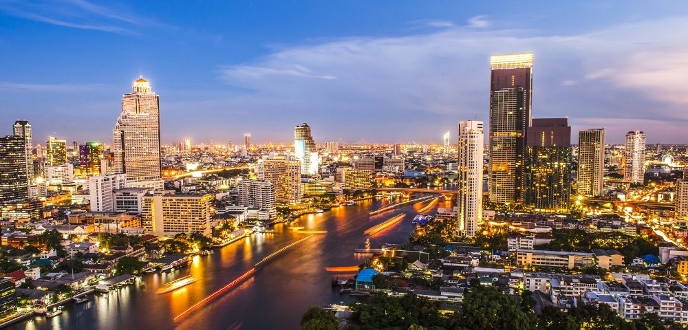 A vibrant cityscape at dusk with illuminated skyscrapers, bustling river traffic, and a twilight sky. The scene conveys energy and urban life.