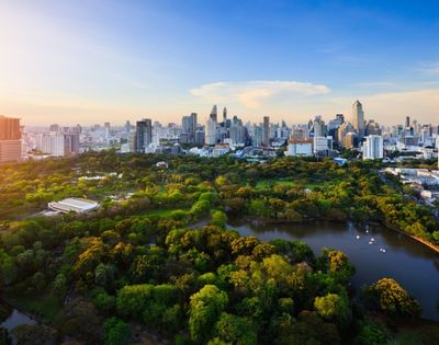Aerial view of a lush, green park with ponds against a backdrop of a bustling city skyline under a bright blue sky at sunrise.