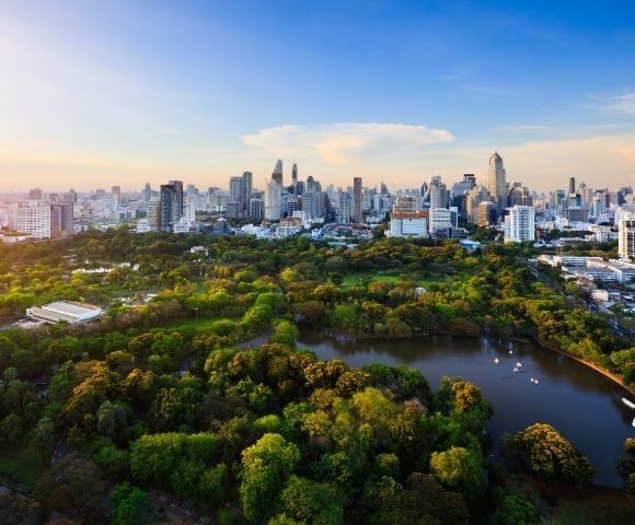 Aerial view of a lush, green park with ponds against a backdrop of a bustling city skyline under a bright blue sky at sunrise.