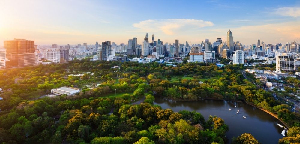 Aerial view of a lush, green park with ponds against a backdrop of a bustling city skyline under a bright blue sky at sunrise.