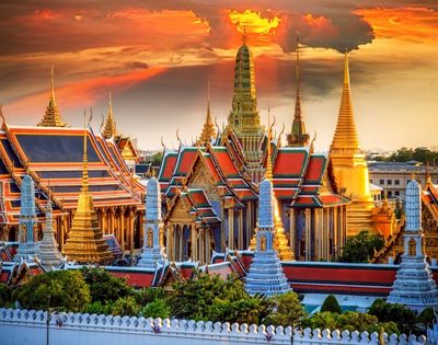 Vibrant sunset over the Grand Palace in Bangkok, featuring ornate golden spires, red and green roofs, and a dramatic sky with orange clouds.
