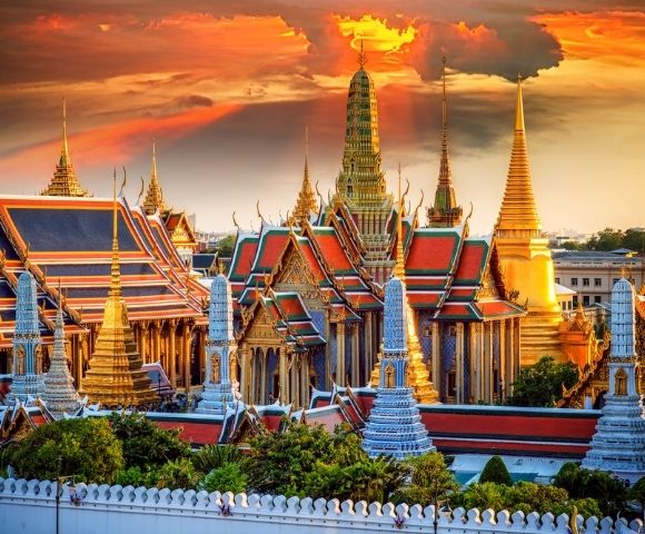 Vibrant sunset over the Grand Palace in Bangkok, featuring ornate golden spires, red and green roofs, and a dramatic sky with orange clouds.
