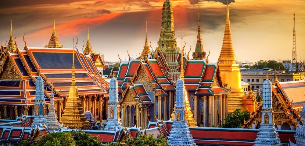 Vibrant sunset over the Grand Palace in Bangkok, featuring ornate golden spires, red and green roofs, and a dramatic sky with orange clouds.