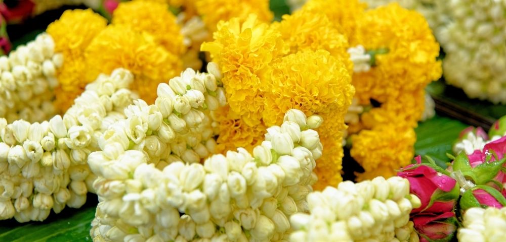 Flower garlands made of yellow marigolds and white jasmine buds are arranged side by side on green leaves, creating a vibrant and festive display.