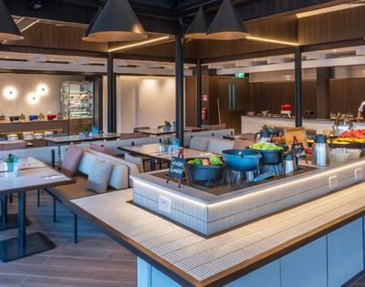 Modern, spacious buffet restaurant with warm lighting, sleek tables, and cushioned seating. Fresh salads and dishes displayed; two chefs visible.