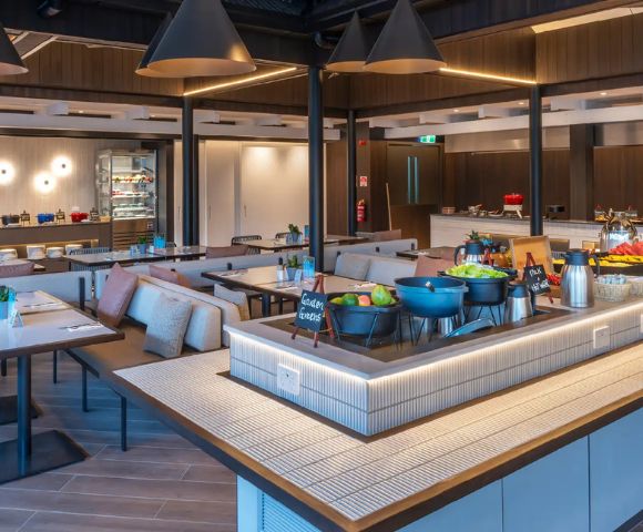 Modern, spacious buffet restaurant with warm lighting, sleek tables, and cushioned seating. Fresh salads and dishes displayed; two chefs visible.