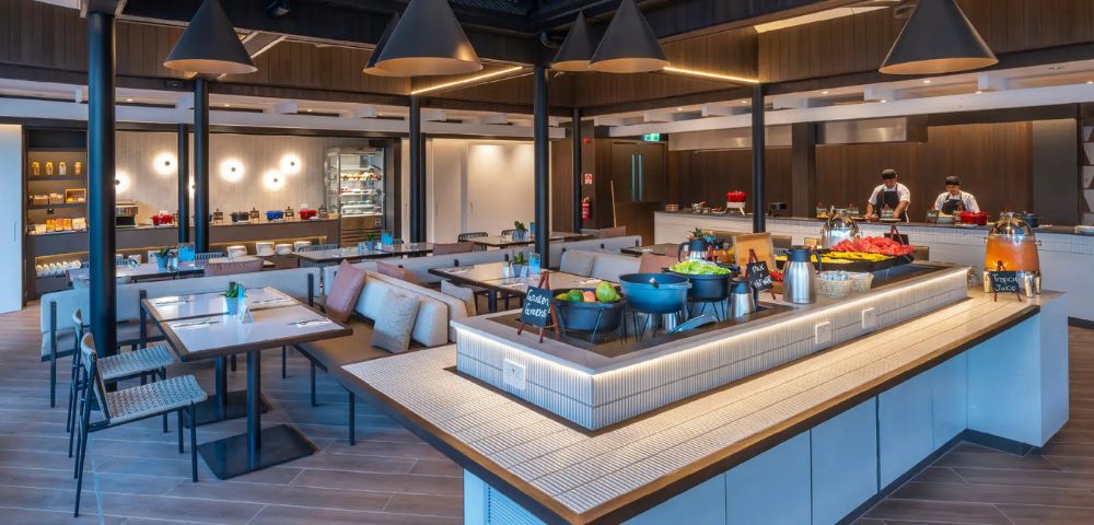 Modern, spacious buffet restaurant with warm lighting, sleek tables, and cushioned seating. Fresh salads and dishes displayed; two chefs visible.