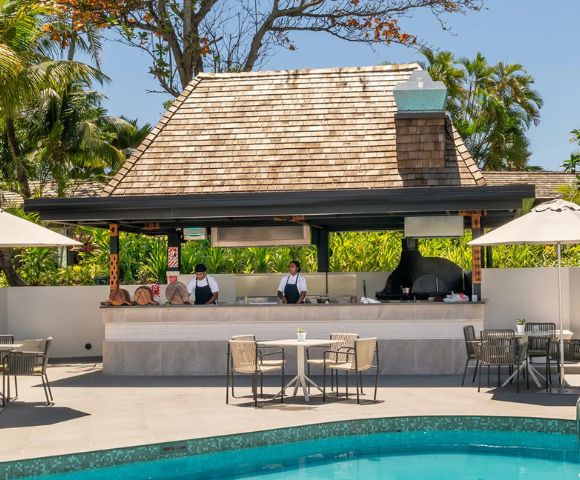 Poolside dining area with a thatched-roof bar, chefs preparing food, and tropical plants. Tables with umbrellas create a relaxed, resort vibe.