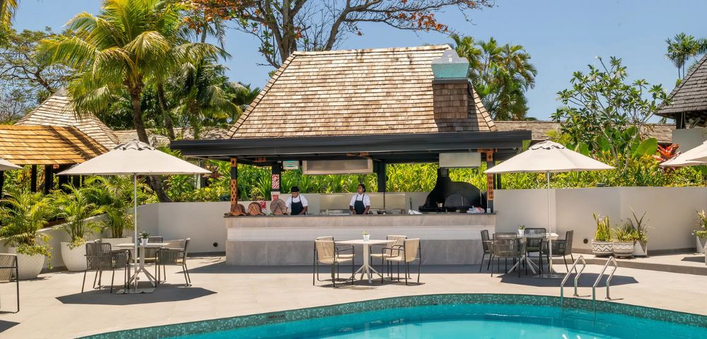 Poolside dining area with a thatched-roof bar, chefs preparing food, and tropical plants. Tables with umbrellas create a relaxed, resort vibe.