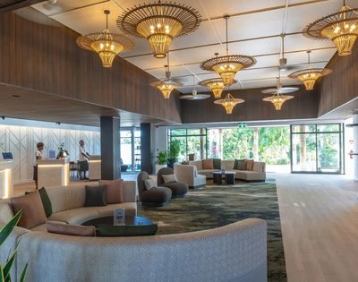 Spacious hotel lobby with elegant wicker chandeliers, plush circular couches, and large windows showing greenery outside. A calm, luxurious atmosphere.