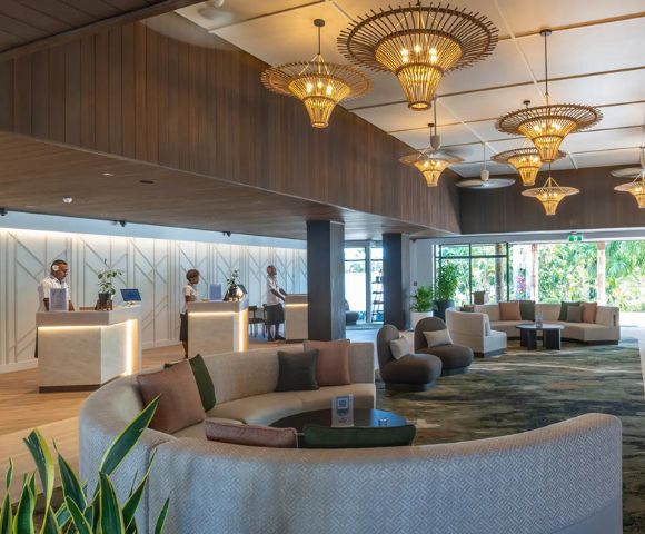 Spacious hotel lobby with elegant wicker chandeliers, plush circular couches, and large windows showing greenery outside. A calm, luxurious atmosphere.