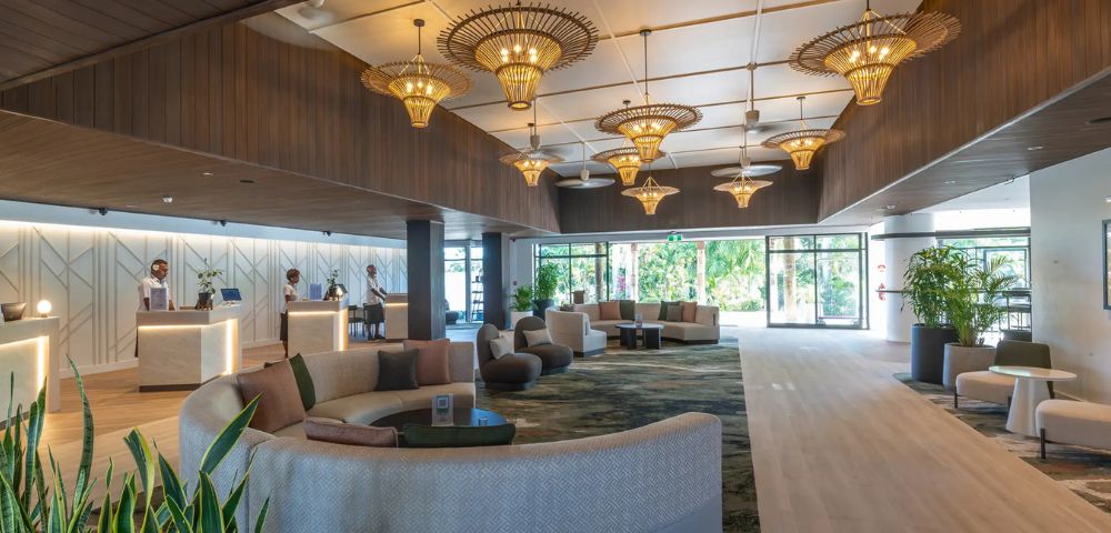 Spacious hotel lobby with elegant wicker chandeliers, plush circular couches, and large windows showing greenery outside. A calm, luxurious atmosphere.