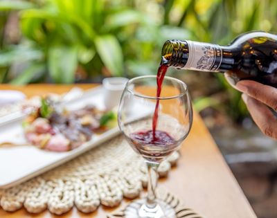 A hand pours red wine into a glass on a wooden table set with a plate of food. Lush greenery in the background creates a fresh, elegant dining atmosphere.