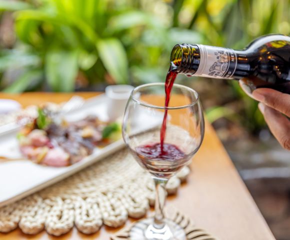 A hand pours red wine into a glass on a wooden table set with a plate of food. Lush greenery in the background creates a fresh, elegant dining atmosphere.
