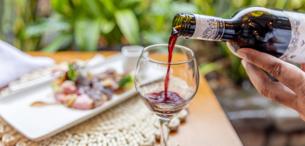 A hand pours red wine into a glass on a wooden table set with a plate of food. Lush greenery in the background creates a fresh, elegant dining atmosphere.