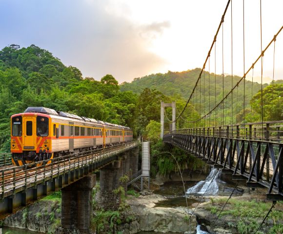 A yellow and orange train crosses a bridge above a lush, green valley with a waterfall. A suspension bridge is on the right, and sunlight filters through the trees.