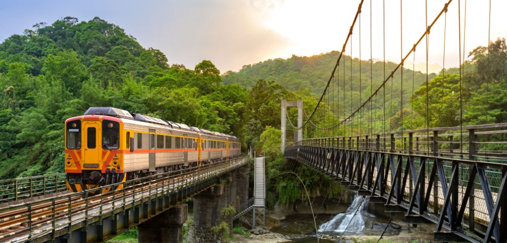 A yellow and orange train crosses a bridge above a lush, green valley with a waterfall. A suspension bridge is on the right, and sunlight filters through the trees.
