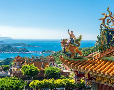 Ornate Taiwanese temple on a sunny day, featuring colorful dragon statues on the roof. Lush greenery surrounds, with blue ocean and islands in the background.
