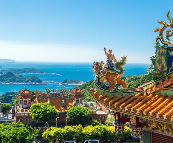 Ornate Taiwanese temple on a sunny day, featuring colorful dragon statues on the roof. Lush greenery surrounds, with blue ocean and islands in the background.