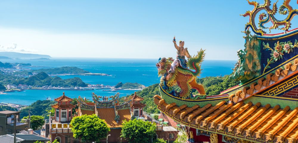 Ornate Taiwanese temple on a sunny day, featuring colorful dragon statues on the roof. Lush greenery surrounds, with blue ocean and islands in the background.