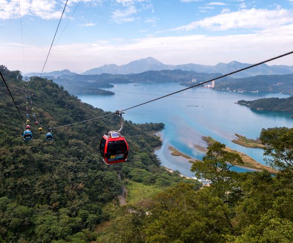Cable cars traverse above a lush forest with a serene lake and mountains in the background, creating a tranquil and scenic aerial view.