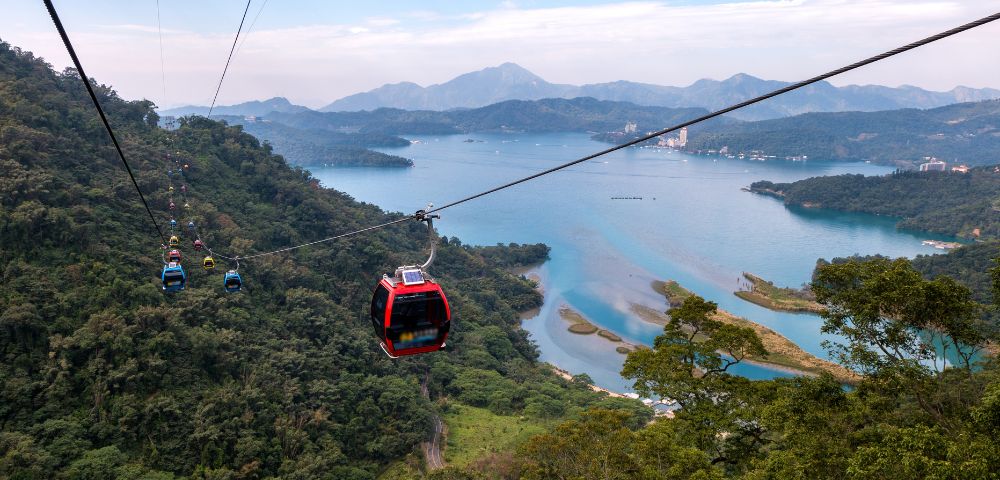 Cable cars traverse above a lush forest with a serene lake and mountains in the background, creating a tranquil and scenic aerial view.