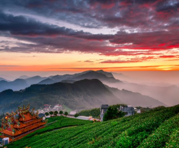 Sunset view of lush green mountains with a majestic temple in the foreground. Dramatic red and orange sky creates a serene and peaceful atmosphere.