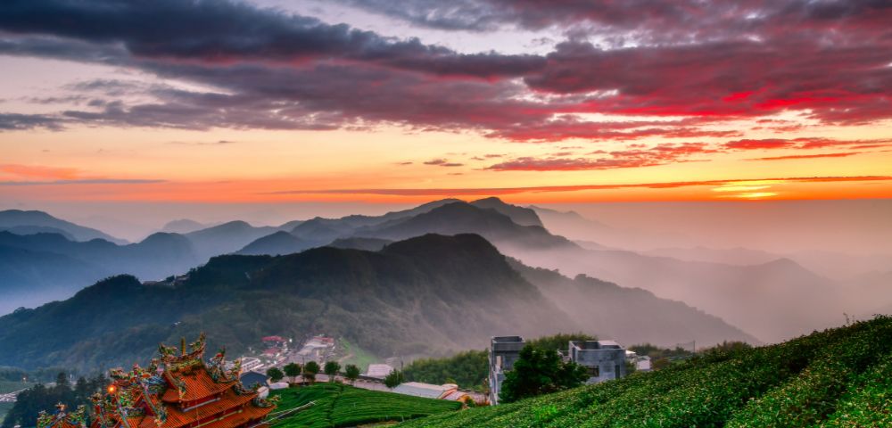 Sunset view of lush green mountains with a majestic temple in the foreground. Dramatic red and orange sky creates a serene and peaceful atmosphere.