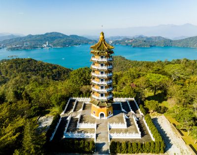 Aerial view of a traditional pagoda surrounded by greenery, set against a backdrop of a serene blue lake and distant mountains under a clear sky.