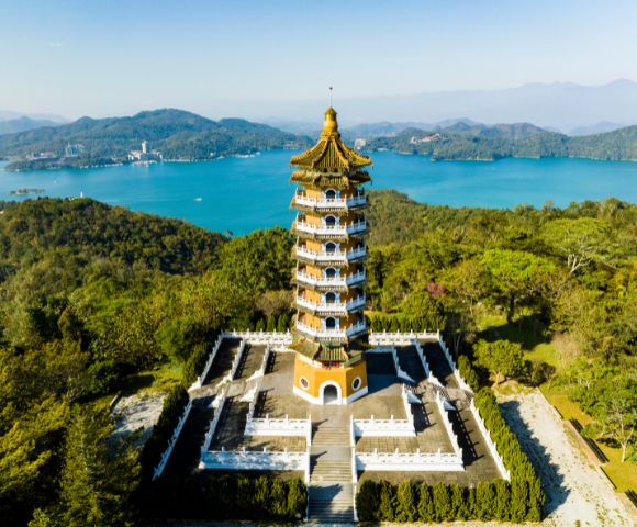 Aerial view of a traditional pagoda surrounded by greenery, set against a backdrop of a serene blue lake and distant mountains under a clear sky.
