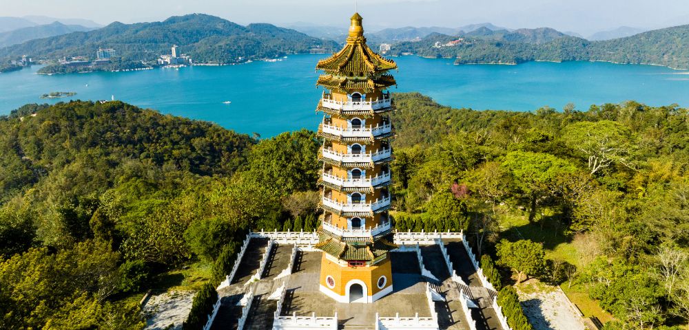 Aerial view of a traditional pagoda surrounded by greenery, set against a backdrop of a serene blue lake and distant mountains under a clear sky.