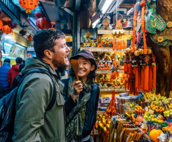 A smiling man and woman enjoy browsing a vibrant street market adorned with colorful lanterns and ornaments, creating a lively and festive atmosphere.