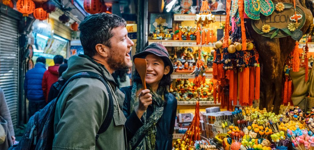 A smiling man and woman enjoy browsing a vibrant street market adorned with colorful lanterns and ornaments, creating a lively and festive atmosphere.