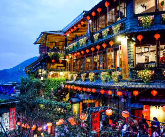 A vibrant, multi-story tea house adorned with glowing red lanterns and string lights, set against a dusky sky. Lush greenery adds charm and warmth.