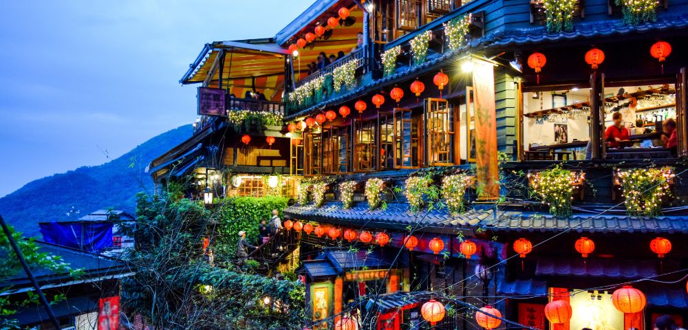 A vibrant, multi-story tea house adorned with glowing red lanterns and string lights, set against a dusky sky. Lush greenery adds charm and warmth.