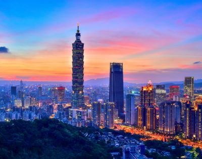 A vibrant cityscape of Taipei at sunset, with Taipei 101 prominently lit against a colorful sky, surrounded by illuminated skyscrapers and a foreground of lush hills.