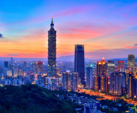 A vibrant cityscape of Taipei at sunset, with Taipei 101 prominently lit against a colorful sky, surrounded by illuminated skyscrapers and a foreground of lush hills.