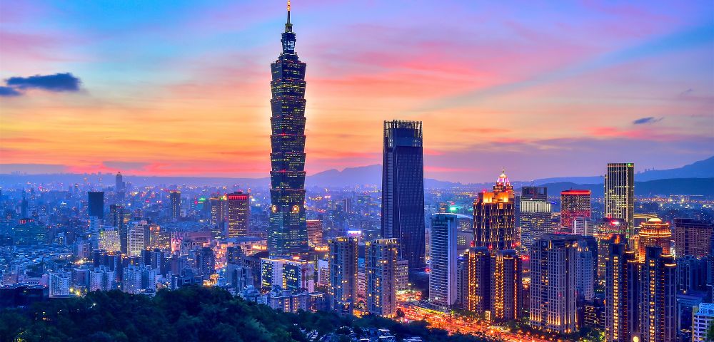 A vibrant cityscape of Taipei at sunset, with Taipei 101 prominently lit against a colorful sky, surrounded by illuminated skyscrapers and a foreground of lush hills.