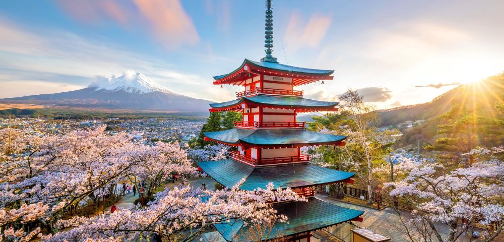 Red pagoda with sloping roofs surrounded by cherry blossoms in full bloom. Mount Fuji and a sunset sky create a serene backdrop.
