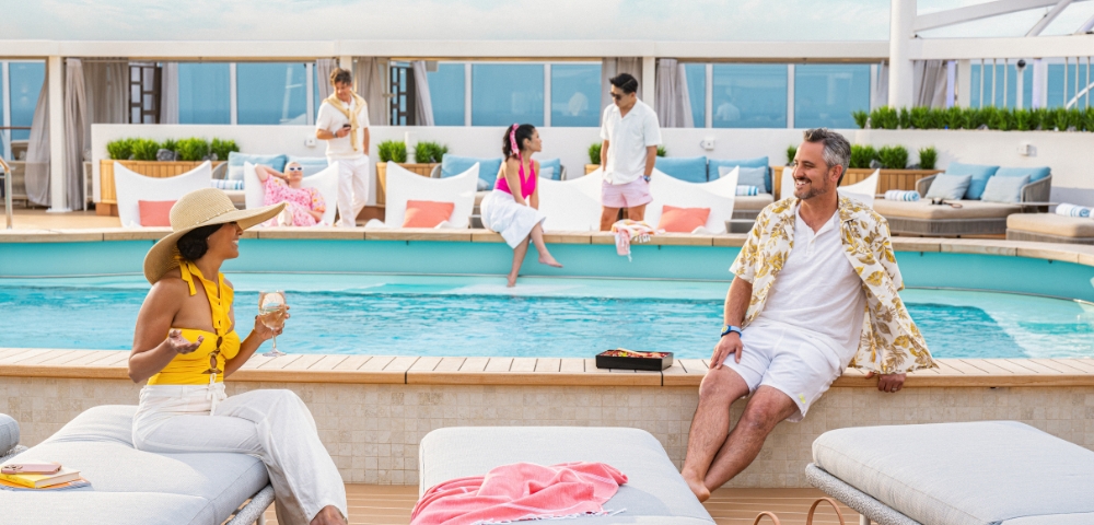 A relaxed poolside scene with adults conversing and smiling. A woman in a hat holds a drink, while others enjoy the sunny day, seated or standing by the pool.
