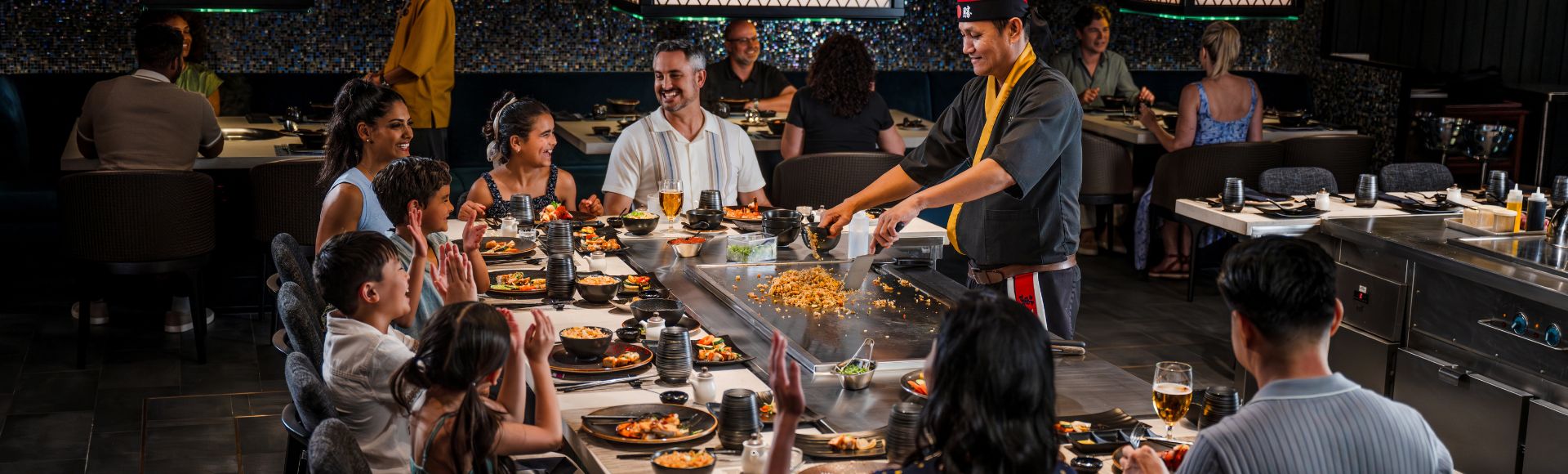 A chef performs a lively teppanyaki cooking show for an attentive, smiling group seated around a hibachi grill in a restaurant.